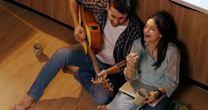 Joyful Indian Asian young couple sitting on their kitchen floor, relaxing and strumming chords on a guitar while singing together in harmony.