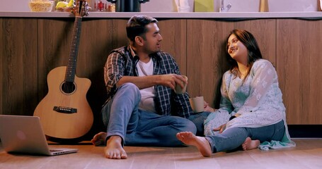 An Indian Asian young couple sitting on the kitchen floor, enjoying coffee together while sharing laughter and moments of joy during their relaxing time