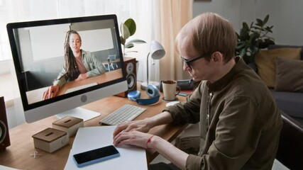 Visually impaired man having video call with young woman on computer, he sitting at wooden desk with braille book