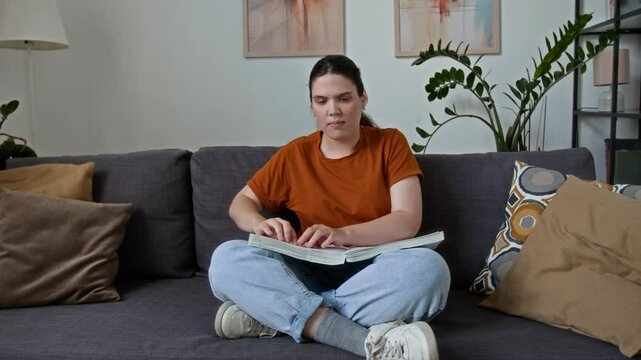 Young woman with disability reading book with braille writing system while sitting on couch at home
