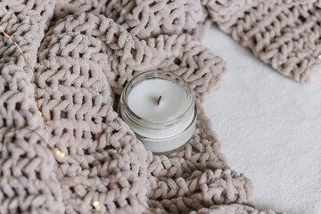 A candle in a glass jar lies among a knitted plush soft warm plaid. Warm comfort