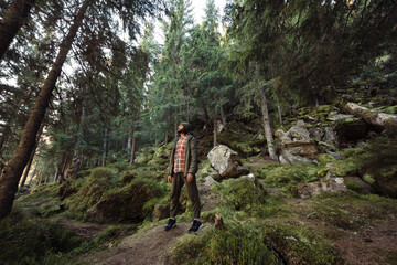 Fototapeta premium A man in casual outdoor attire stands amidst a lush forest, surrounded by towering trees and moss-covered rocks. Ideal for high-quality content focusing on nature adventure, hiking, and outdoor