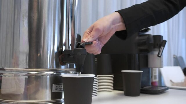 A woman's hand takes a disposable cup and pours hot water into it. Self-service or making tea or coffee in the office.