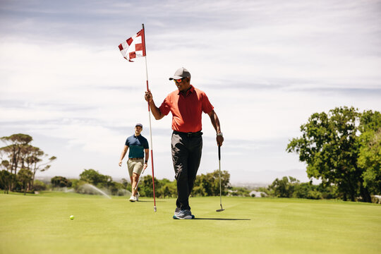 Golfers walking on putting green, holding golf clubs and flagstick
