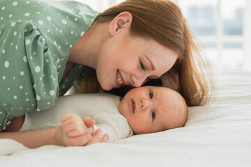 Happy family at home. Mother playing with little toddler child on bed. Mom baby relax playing having fun together. Mother looks at baby with love care. Mom of breast feeding baby rest in living room