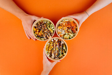 Hands holding vibrant poke bowls on orange background, celebrating healthy eating and shared meals