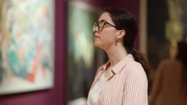 Side view of young Caucasian beautiful woman with glasses watching an exhibition of paintings in a gallery. The concept of cultural education.