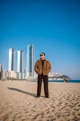 A Korean guy in a leopard fur coat walks along the beach against the backdrop of the autumn landscape of Busan Haeundae.