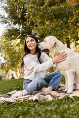 Fototapeta premium A young woman in cozy attire joyfully interacts with her dog in a sunlit park.