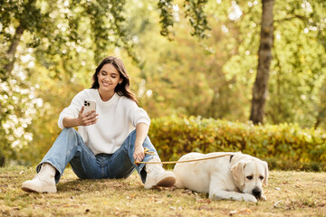 Fototapeta premium A beautiful young woman relaxes in the park, checking her phone beside her dog on a warm autumn day.