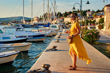 Woman is walking alone on sunny promenade with palm trees and flowers. Female tourist strolls along marina at sunset in coastal town © Lazy_Bear