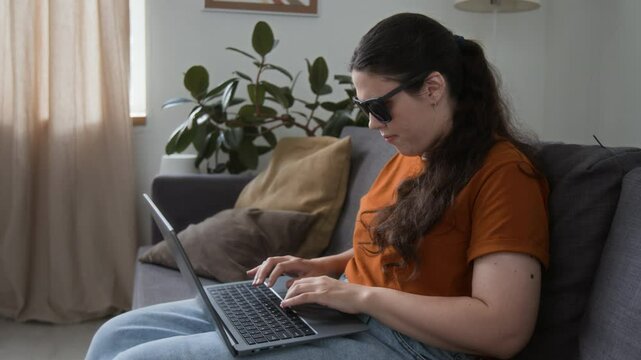 Visually challenged young woman with black glasses working on laptop while sitting on couch