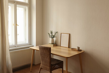Empty vertical picture frame mockup. Wooden desk, table. Vase with olive branches, books, cup of coffee. Elegant working space, home office. Summer interior design. Beige walls. Blurred curtain window