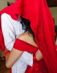Initiation procedure, a man in white and a woman in a red dress, standing embracing each other under a red cloth; visible body parts.