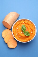 Tasty mashed sweet potato with basil, seeds in bowl and cut vegetable on blue background, flat lay
