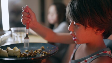 Fototapeta premium Young boy eating at the dining table, holding a fork and focusing on the food on his plate during meal time in a relaxed home setting