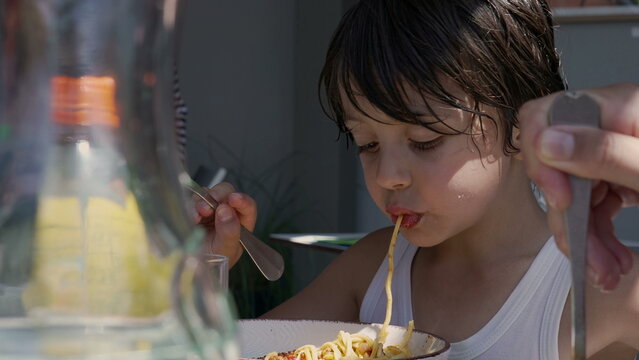 Young boy slurping a long noodle while eating spaghetti, using a fork to twirl the pasta and enjoying his meal outdoors at a table during the day