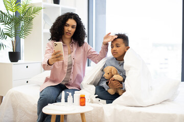 Concerned African mother checks temperature of sick young boy while using phone for online doctor consultation. Boy, wrapped in blanket, holds teddy bear on bed, surrounded by medicine.