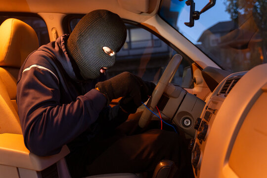 Masked thief hotwiring car ignition system in vehicle theft attempt