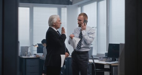 Serious professional stock trader, stockbroker speaks with business client on call using mobile phone. Senior boss of the company stands near and listens to conversation. Work in modern office.