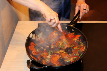 Cooking a delicious dinner for your family in the kitchen in a frying pan (roast with meat and vegetables, mushrooms, red pepper, chili garlic and others). The dish is steaming