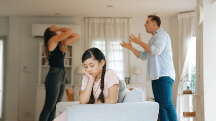 Annoyed and unhappy young girl sitting on sofa trapped in middle of tension by her parent argument in living room. Unhealthy domestic lifestyle and traumatic childhood develop to depression Synchronos