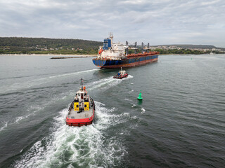 Aerial view of Tug boats assisting big cargo ship. Large cargo ship enters the port escorted by...