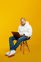 Vertical photo of full body young man sitting on chair and using modern notebook, typing on laptop, isolated on yellow background