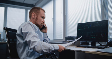 Analyst studying papers with reporting information and discussing results of sales department with colleague on mobile phone. Financial indicators and diagrams displayed on computer screen. Business.