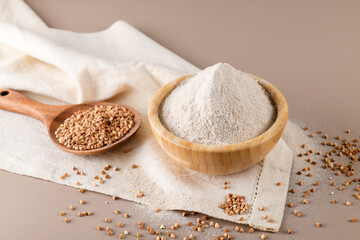 Buckwheat flour in a wooden bowl and raw buckwheat grain on cotton napkin on beige monochrome background. Alternative flour, gluten free flour, healthy nutrition