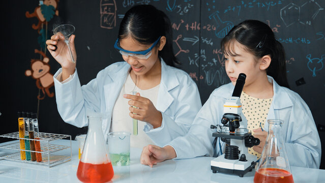 Young highschool student doing experiment by pouring sample in test tube. Girl wearing glasses and looking under microscope to inspect sample in STEM science class. Creative education. Edification.