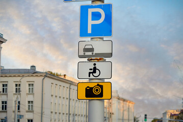 Parking sign on pole with accessibility and surveillance camera symbols in an urban environment, traffic regulation, security measures, and designated disabled parking areas in the city.