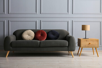 Stylish sofa with cushions and lamp on side table near grey wall