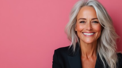 A sophisticated woman with silver hair and a warm smile is captured standing against a pink backdrop, embodying timeless elegance and charm in her black attire.