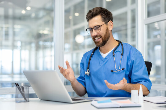 Smiling doctor wearing blue medical uniform conducts online consultation using laptop in modern office. Stethoscope around neck, bright windowed background, conveying professionalism.