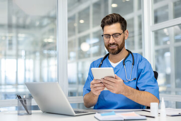 Healthcare professional in scrubs using smartphone in modern office. Man sits at desk with laptop,...