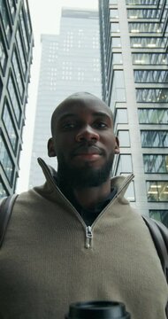 A man in a sweater, with a briefcase on his shoulders and a disposable cup of coffee in his hand looks at the camera with a calm face standing on a city street