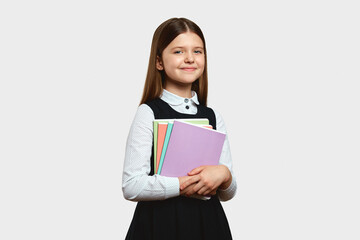 Cheerful pupil girl with blonde hair wearing school uniform, embracing colorful notebooks and looking at camera, isolated over white background