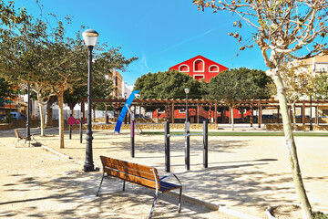 Urban park area with wooden bench next to black lamppost, fitness or workout area, surrounding with neatly pruned trees. Outdoor activities or relaxation. Costa Blanca, Almoradi town, Spain

