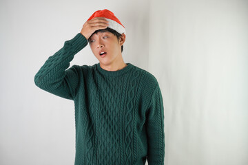Sick young asian man wearing santa clause hat and green long sleeved sweater is suffering headache, isolated over white background. Concept for Christmas Holiday and New Year Party