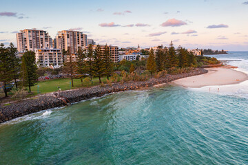 Aerial view of high rise buildings and a coastal reserve above a rocky breakwall and beach