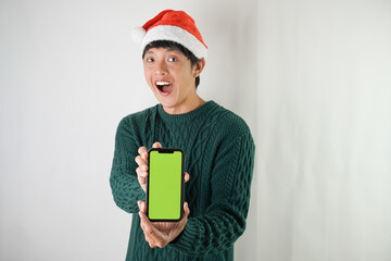 Excited young asian man wearing santa clause hat and green long sleeved sweater is showing smartphone green screen, isolated over white background. Concept for Christmas Holiday and New Year Party