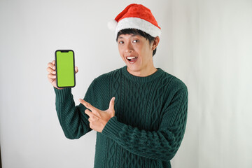 Excited young asian man wearing santa clause hat and green long sleeved sweater is showing smartphone green screen, isolated over white background. Concept for Christmas Holiday and New Year Party