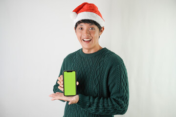 Excited young asian man wearing santa clause hat and green long sleeved sweater is showing smartphone green screen, isolated over white background. Concept for Christmas Holiday and New Year Party