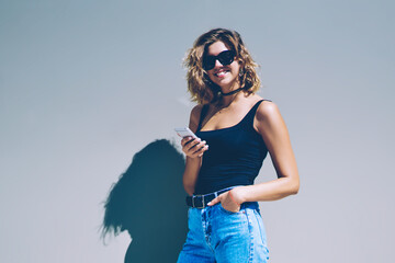 Full length portrait of cheerful hipster girl with smartphone in hand and looking at camera on urban setting, happy woman in sunglasses browsing internet on cellular phone during sunny summer day