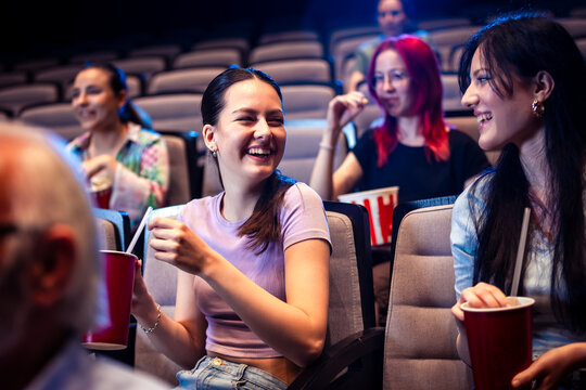 Two best friends are watching a movie in cinema and they are eating popcorn.