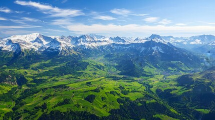 Panoramic view of snow-capped mountains with green valleys and meadows under a blue sky with clouds.