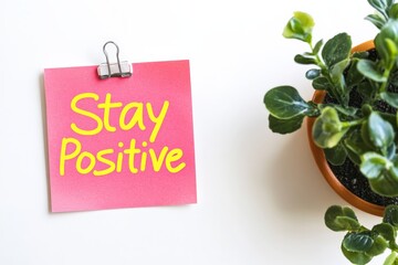 Featuring 'Stay Positive' in vivid yellow on a pink note with a binder clip, this image includes a green potted plant, blending optimism and nature's calm essence.