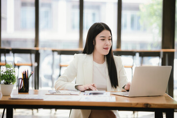 Beautiful Asian female accountant working with smartphone and laptop at her desk analyzing business reports and documents. Sending messages