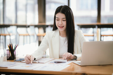 Beautiful Asian female accountant working with smartphone and laptop at her desk analyzing business reports and documents. Sending messages